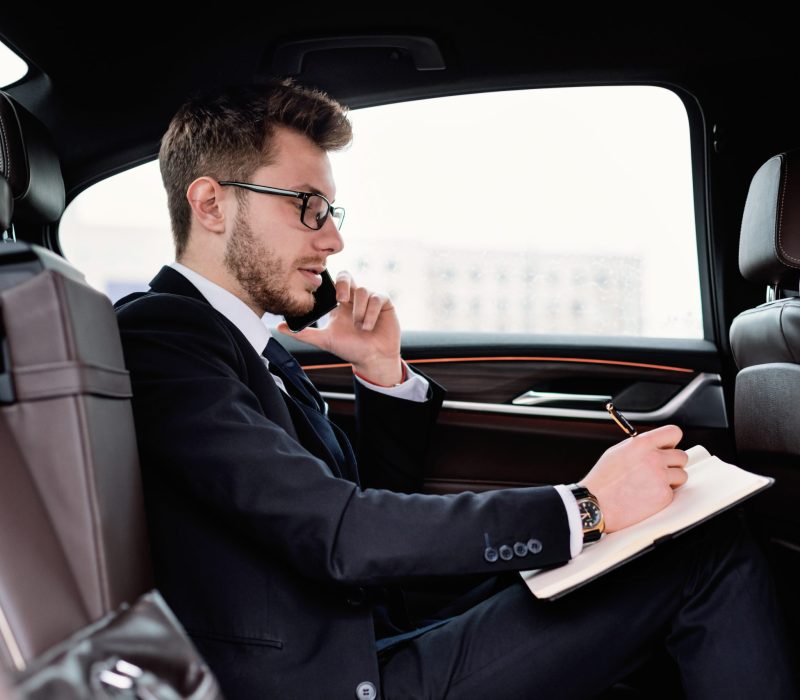 Test Drive Concept. Young manager in glasses writing in notebook with pen sitting in back seat of new car, side view