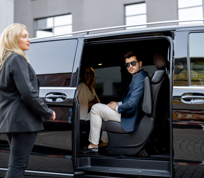 Female chauffeur helps a business man to get out of vehicle, opening a door of luxury black minivan. Concept of personal driver, luxury taxi for business delegation